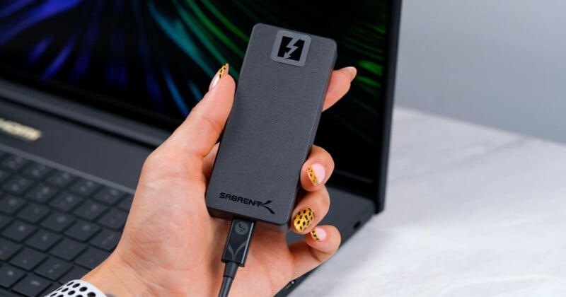 A hand with decorated nails holds a black Sabrent external SSD, which has a lightning bolt icon. The SSD is connected to a laptop in the background.