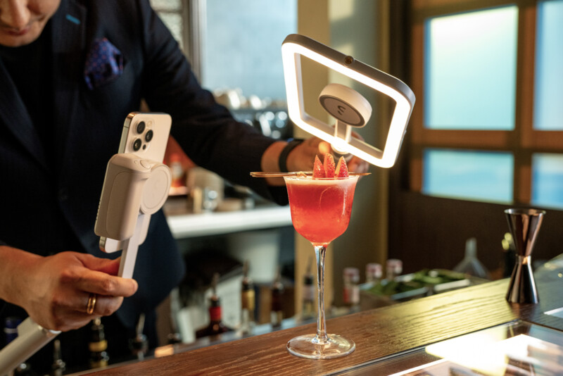 A person is setting up a smartphone on a gimbal with a ring light to take a photo of a vibrant red cocktail garnished with citrus zest. The cocktail is placed on a wooden bar countertop with various bar tools and bottles visible in the background.