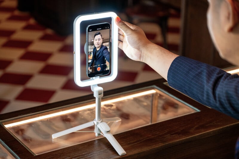 A person is holding a smartphone mounted on a tripod with a ring light. The screen shows the individual taking a selfie in a well-lit indoor setting. The floor has red and white checkered tiles and the person is wearing a dark outfit.