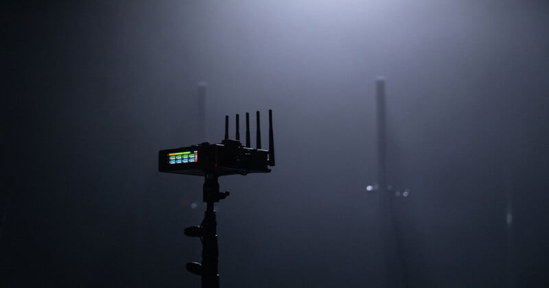 A small electronic device with antennas is mounted on a stand, illuminated by soft overhead lighting. The background is dark and blurry, giving a mysterious atmosphere. Colored lights can be seen on the device's display.