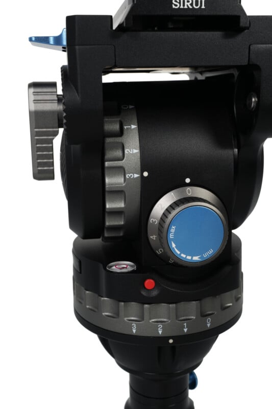 Close-up of a black and silver camera tripod head with blue adjustment knobs and various numerical settings. The tripod head is labeled as "Sirui" and has intricate gears and a red button for precise adjustments.