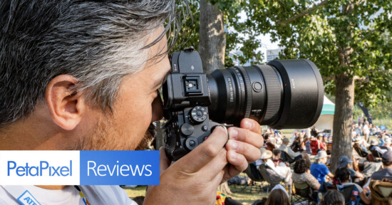 A person with short gray hair is closely holding and looking through a professional camera. The scene is outdoors with trees and a crowd in the background. The image has banners with the text "PetaPixel" and "Reviews.