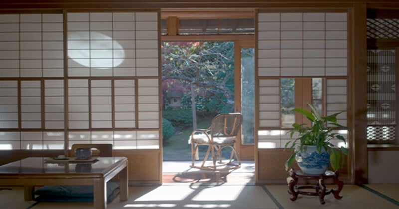 A serene Japanese room with shoji screens partially open, allowing a view of a garden with a tree. Light creates a circular shadow on the screens. Inside are a low wooden table with tea items and a woven chair by the doorway. A potted plant rests on a stand.