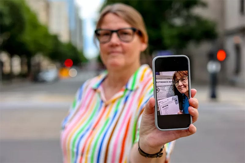 A person wearing glasses and a colorful striped shirt holds a smartphone with their other hand. The phone screen shows a close-up of the person smiling and holding a document. The background is an urban street scene with trees and buildings out of focus.