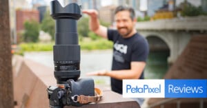 A camera with a large lens is placed on a ledge outdoors, with an arm strap attached. In the blurred background, a smiling man stands next to a river and bridge, holding his hands out as if framing the shot. The image is overlaid with the logo and text "PetaPixel Reviews.