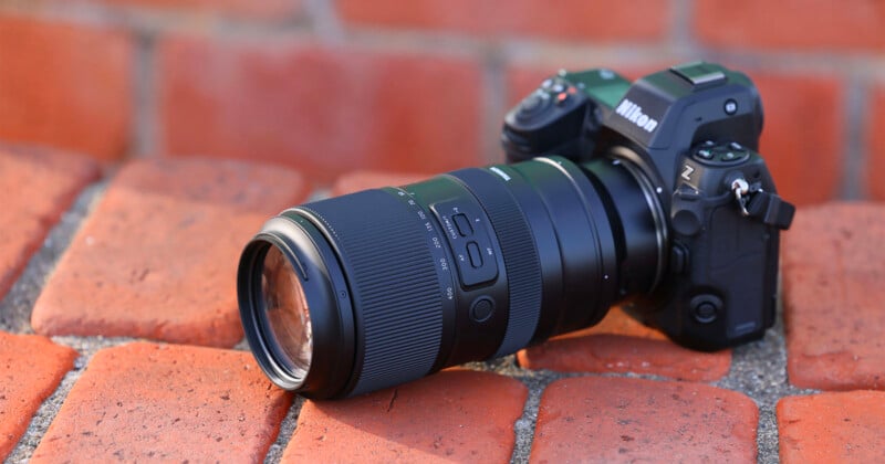 A Nikon camera with a large zoom lens attached lies on its side on a surface made of red bricks. The camera has a black body and multiple buttons and dials. The lens has grip texture and switches for different settings.