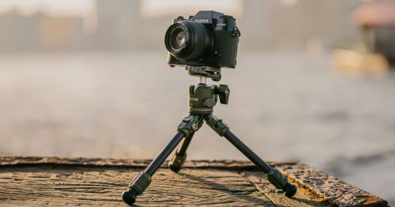 A black Fujifilm camera mounted on a compact tripod is placed on a wooden surface. The background is blurred, showing an out-of-focus waterfront with buildings, suggesting an outdoor setting during golden hour.