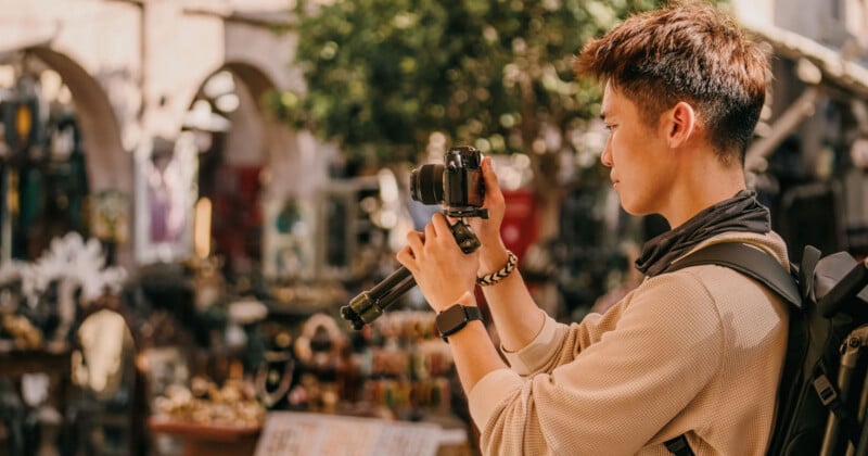 A person with short hair and a backpack is taking a photo with a camera on a tripod. They are standing in an outdoor market area with various items and stalls in the background, including trees and archways. The sun is shining, and the atmosphere looks warm and vibrant.