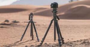 Two cameras mounted on tripods stand in a desert landscape with sand dunes in the background. The cameras are set up at different distances, capturing the arid scenery under a clear sky.