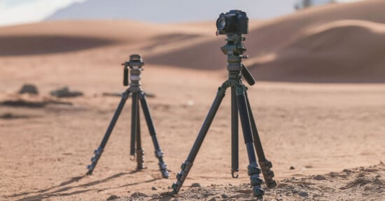 Two cameras mounted on tripods stand in a desert landscape with sand dunes in the background. The cameras are set up at different distances, capturing the arid scenery under a clear sky.