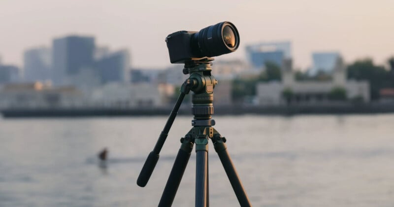 A digital camera mounted on a tripod stands in focus overlooking a body of water and a blurry cityscape in the background during sunset. One person is visible swimming in the water in the distance.