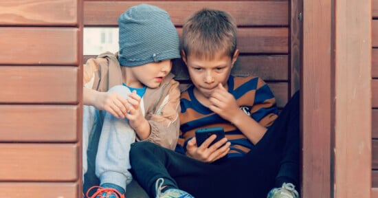 Two young boys sit closely together in a wooden structure. One boy, wearing a striped beanie and light blue sneakers, is looking intently at a smartphone held by the boy next to him, who is wearing a striped shirt and black pants. Both appear focused on the screen.