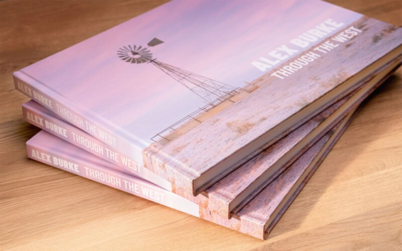 A stack of hardcover books titled "Alex Burke: Through the West" is shown on a wooden surface. The cover features a scenic photograph of a windmill on a rural landscape under a soft, pastel sky.