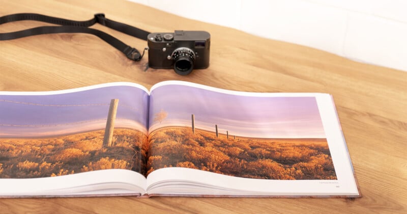 An open photography book displaying a landscape image of a rural scene with a fence and golden foliage. A black camera with a strap is placed on a wooden table in the background. The image has a warm and tranquil ambiance.