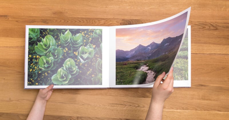 Hands holding a photo book open on a wooden table. The left page displays close-up images of green cacti with yellow flowers, and the right page shows a scenic mountain landscape with a winding path and a vibrant sunset sky.