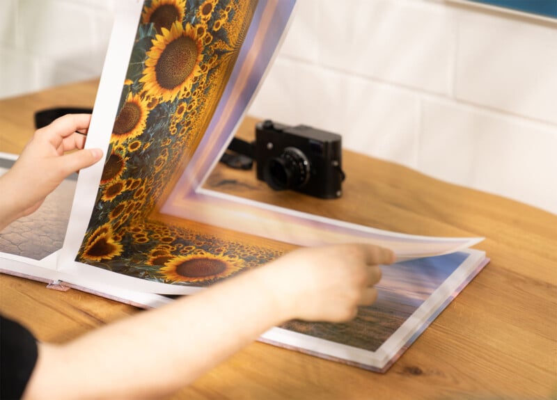 A person flipping through a large photo book with images of sunflowers on a wooden table. There is a black camera placed on the table in the background, and the setting appears to be indoors with white tiled walls.