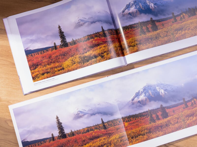 Two open landscape photography books are laid out on a wooden surface. Both books feature a stunning autumnal scene with vibrant red, orange, and yellow foliage in the foreground, snow-covered mountains, and a partly cloudy sky in the background.