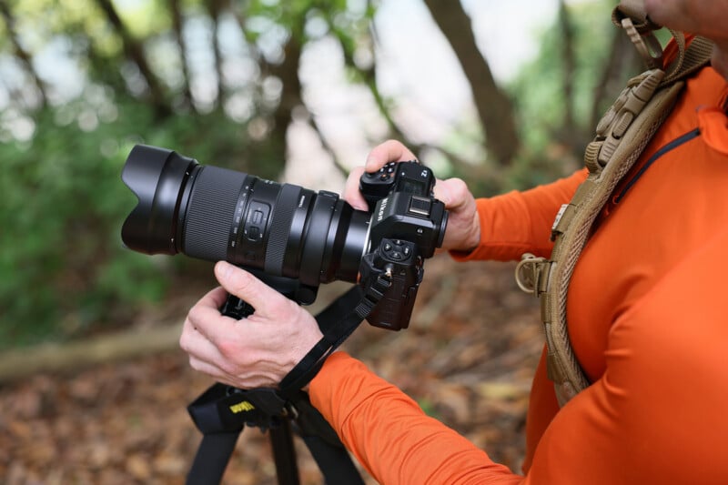 A person wearing an orange long-sleeve shirt holds a DSLR camera with a large lens attached, mounted on a tripod. The background features blurred greenery and tree trunks.