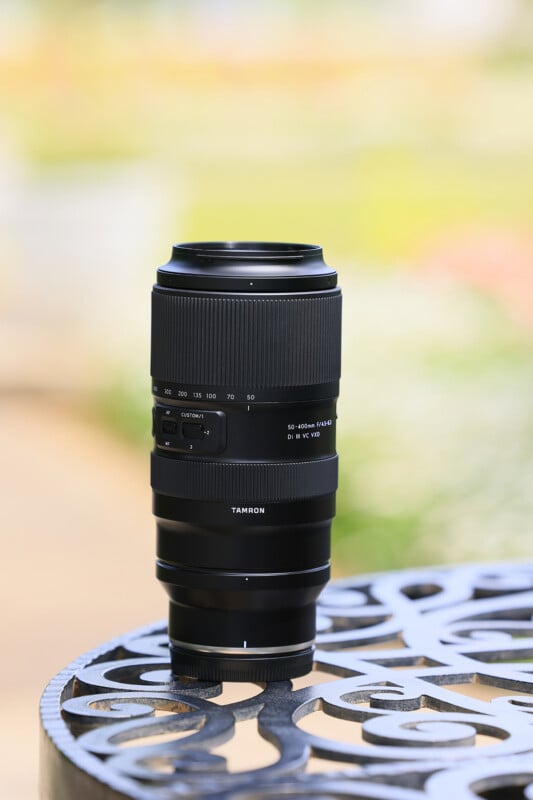 A black Tamron 50-400mm camera lens with white text detailing sits upright on an ornate metal table. The background is slightly blurred, featuring light and soft pastel colors, suggesting an outdoor setting in a garden or park.