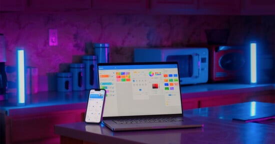 A laptop and smartphone displaying smart home apps are placed on a counter in a kitchen with colorful ambient lighting. LED light bars on either side of the counter emit blue and purple hues, enhancing the modern and high-tech atmosphere of the room.