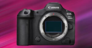 A Canon EOS R5 Mark II camera without a lens, showcasing the camera body and sensor. It has a textured grip on the left and various buttons on the top and front. The background features a gradient of pink and purple hues.