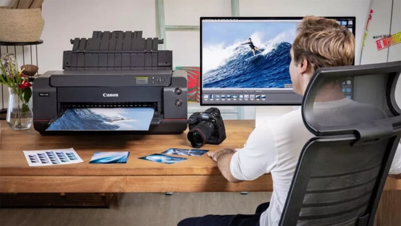 A man sits at a wooden desk using a desktop computer and a Canon printer. Photographs of waves are scattered on the desk and a digital image of a surfer is displayed on the computer monitor. A camera is placed beside the man, who appears focused on his work.