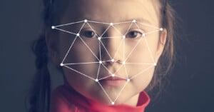 A close-up of a child's face with a digital facial recognition grid overlay. The grid highlights key points and lines across the face, including the eyes, nose, and mouth. The child has a neutral expression and is wearing a red top, with dark background.