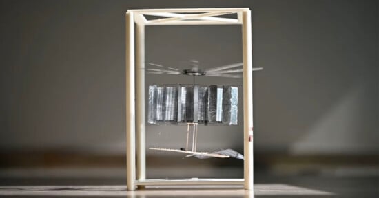 A small, complex mechanical device with rotating blades is encased in a minimalistic rectangular frame. The backdrop is a simple, blurred gray surface, emphasizing the intricacies and motion of the gadget within the frame.