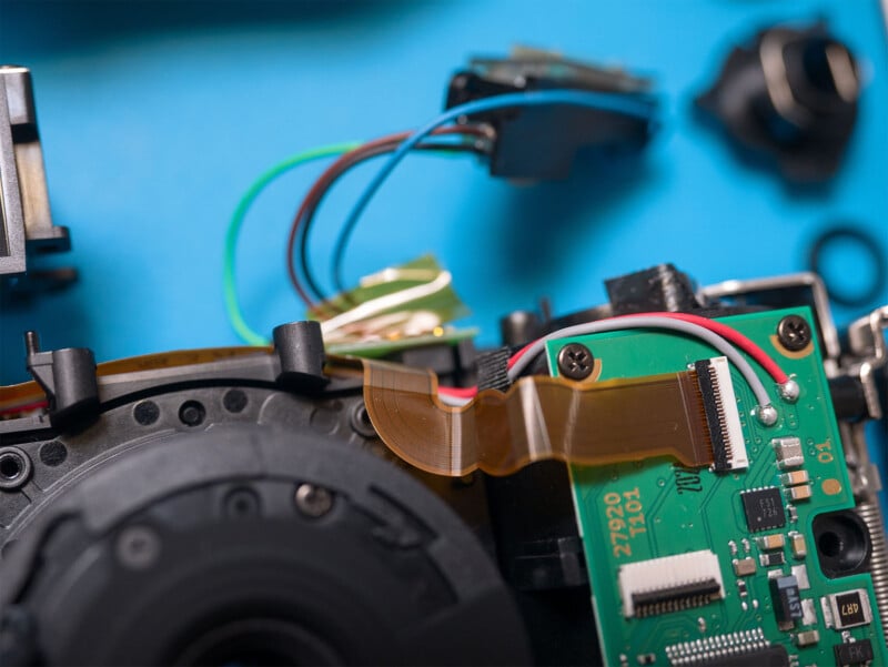 Close-up view of the internal electronics of a disassembled camera on a blue surface, featuring various circuit boards, wires, connectors, and a ribbon cable. Some components are scattered around, indicating a repair or deconstruction process.