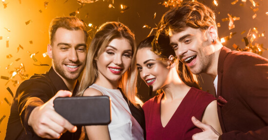 Four people are taking a selfie together at a celebration, smiling and appearing joyful. The background is filled with golden confetti and warm lighting, creating a festive atmosphere.