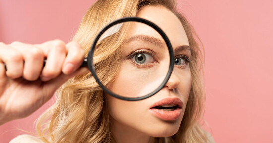 A woman with blonde hair holds a magnifying glass up to her face, which enlarges her right eye. She has a surprised expression and a pink background behind her.
