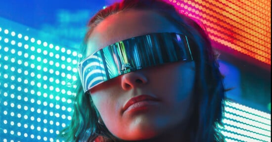 A person with dark shoulder-length hair wears an oversized futuristic visor that covers their eyes. The background is illuminated with vibrant neon lights in blue, pink, and red hues, contributing to a high-tech, cyberpunk atmosphere.