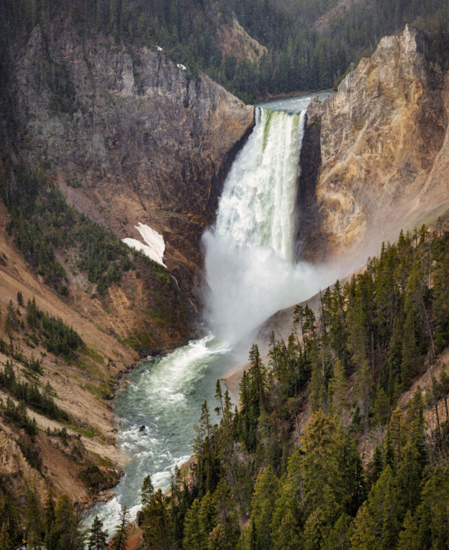 A majestic waterfall cascades down a rocky cliff surrounded by lush green trees and rugged terrain. The water plunges into a river that winds through a forested canyon. Mist from the fall rises into the air, adding a mystical touch to the scene.