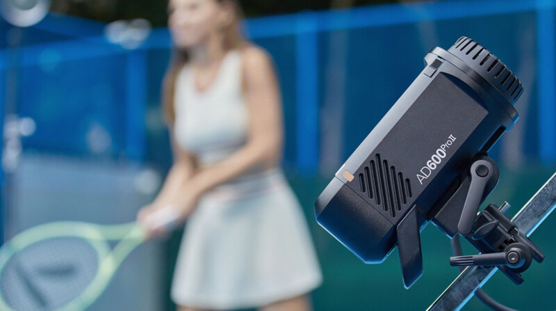 A close-up shot of a black AD600Pro flash unit mounted on a metal support. In the blurred background, a person in a white outfit is preparing to hit a tennis ball with a racket on a tennis court. Blue fencing surrounds the court.