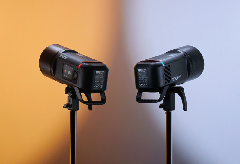 Two black professional studio lights, one labeled Godox and the other labeled Aputure, are mounted on stands against a bi-colored background of orange and blue. Both lights have their controls and labels visible, displaying technical details and certification marks.