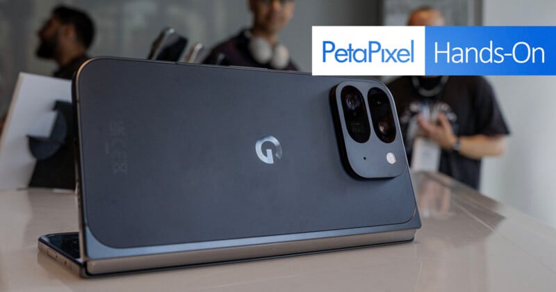 A modern smartphone with a sleek design is displayed on a reflective surface. The back of the phone has a camera module with three lenses shaped like a rectangle. In the background, a blurred image of two people can be seen. The text overlay reads "PetaPixel Hands-On.