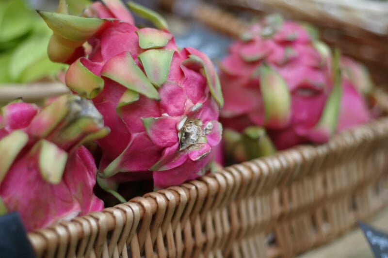 Close-up of a wicker basket filled with bright pink dragon fruits, showcasing their spiky green-tipped scales. A few green leaves are visible in the background, slightly out of focus. The dragon fruits' vibrant colors stand out against the neutral-toned basket.