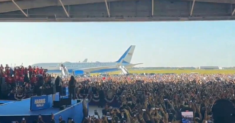 A large crowd gathers in an open hangar for a political rally. The stage displays a "Harris Wally" sign. Two large aircraft, including one with "United States of America" marked on it, are parked in the background against a clear sky. Many people are taking photos.