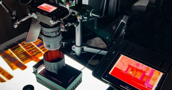 A close-up view of a professional film scanner setup, with a camera and various attachments positioned over film strips on a light table. A tablet displays the digitized images from the film, showing a screen with vibrant colors.