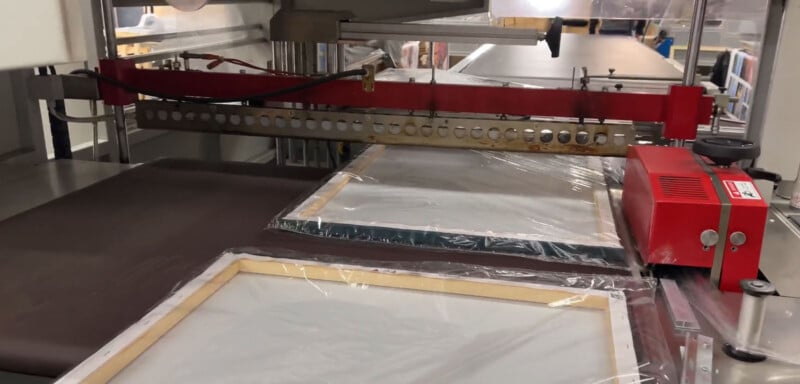 A screen-printing machine in operation, with printing frames under a red bar. The frames are covered in mesh screens, and a brown fabric is moving through the device. The background shows other printing equipment and machinery.