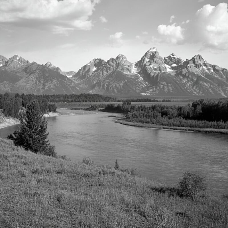 A serene black and white landscape photograph featuring a winding river flowing through a forested area, with a range of snow-capped mountains towering in the background against a partly cloudy sky.