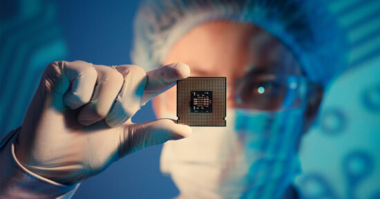 A person in sterile lab attire, including gloves, a mask, and a hairnet, holds a microchip carefully between their thumb and forefinger. The background features a soft, blurred blue circuit pattern.