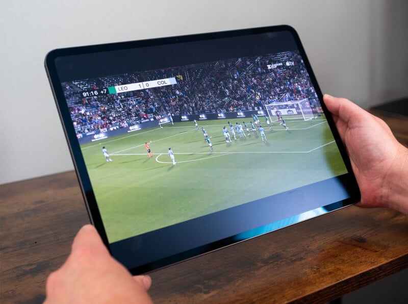 A person is holding a tablet displaying a soccer match. The screen shows a set piece situation with players from two teams on the field, one wearing white and the other in light blue. The score is 1-0, and the timer reads 91:16 with 7 minutes of stoppage time.