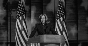 A woman stands at a podium, smiling and speaking into multiple microphones. Two U.S. flags are displayed behind her. The photo is in black and white, giving the scene a formal and historic atmosphere.