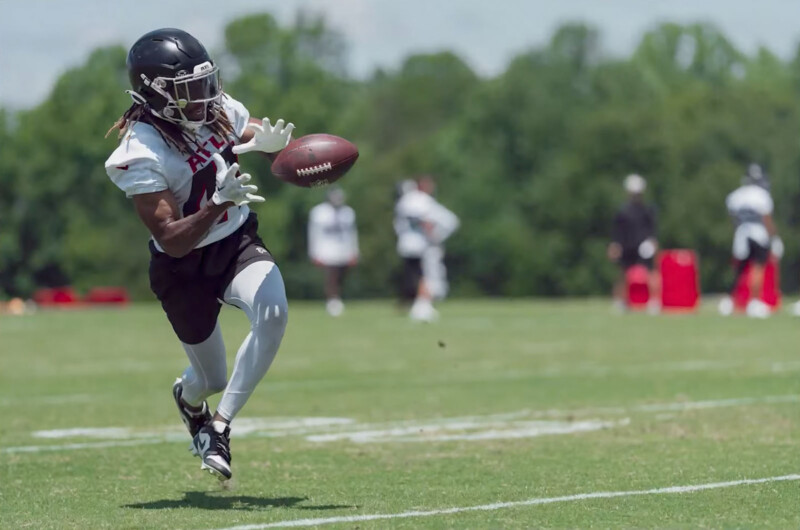 A football player wearing a black helmet, white jersey, and black shorts is catching a football mid-air during a practice session on a green field. Other players and a coach are visible in the background, with trees providing a natural backdrop.