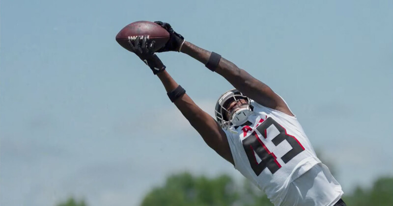 A football player in a white jersey with the number 43 leaps into the air with outstretched arms to catch the football. The sky is clear blue, and green trees are visible in the background.