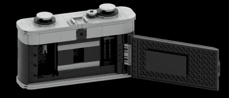 A black and gray LEGO replica of an old-fashioned film camera with the back cover open, showing the internal compartments where film would be loaded. The camera features two knobs on top for adjusting settings. The background is solid black.