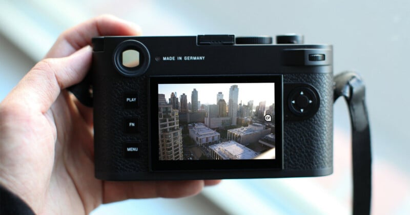 A hand holds a black digital camera with visible controls such as "Play," "Fn," and "Menu." The camera screen displays a cityscape with tall buildings under a clear sky. The camera has a textured grip, and the label "Made in Germany" is visible above the screen.