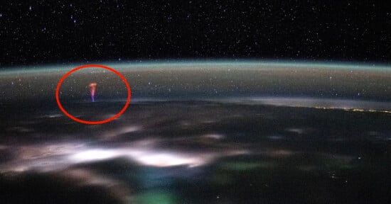 A nighttime view from space showing the curvature of Earth, a brilliant display of stars, and a red sprite lightning phenomenon circled in red near the horizon. The image captures the contrast between the planet's darkness and the outer atmosphere's glow.