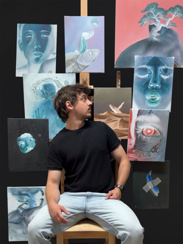 A man with light brown hair and a short beard sits on a wooden stool, wearing a black t-shirt and light blue jeans. He gazes to the side while posing in front of a display of various expressive paintings featuring surreal and abstract themes, all mounted on a black backdrop.